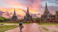 A woman holding a hat in her head stands in front of Wat Phra Si Sanphet Buddhist temple in Thailand